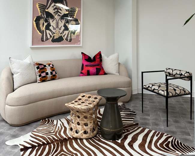 A modern living space features a curved beige sofa with colorful pillows, a round black table, and a patterned chair, all situated on a zebra-striped rug beneath a glass ceiling.