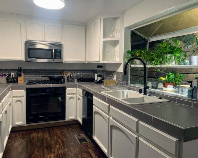 A modern kitchen with white cabinetry, gray tiled backsplash, and a window overlooking a small garden, featuring a sink and stainless steel appliances.