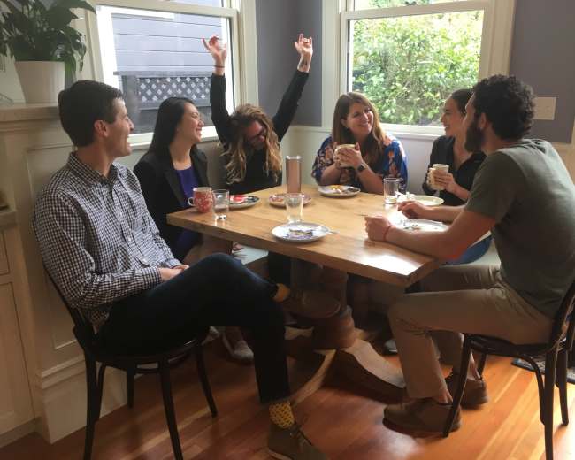 A group of six people sits around a table in a well-lit room, engaging in conversation while enjoying food and drinks.