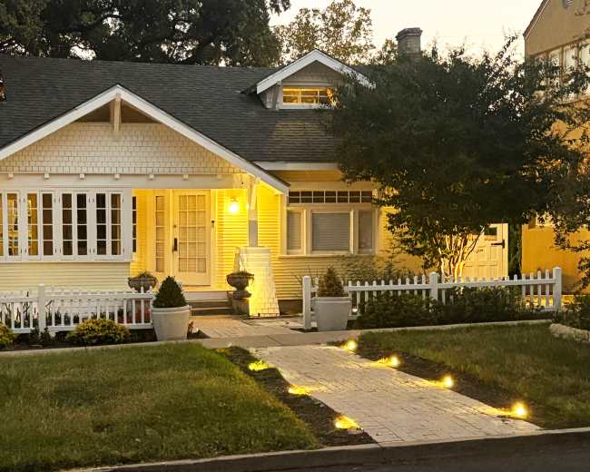 A well-lit, charming bungalow with a white picket fence and garden path lined with lights.