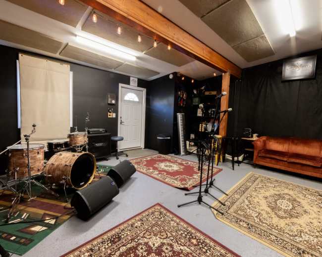 A music rehearsal space features a set of drums, microphones on stands, a keyboard, and a brown couch, all set against dark walls and a door leading outside.