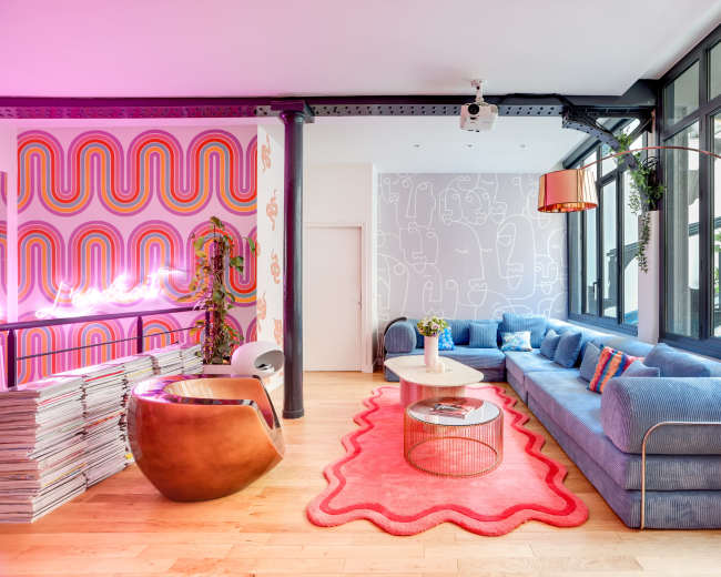 The image shows a modern lounge area featuring a blue sectional sofa, a round coffee table, a curved armchair, and colorful wall decorations.