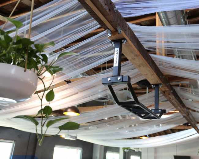 The image shows a black overhead pull-up bar mounted to wooden beams in a room with sheer white drapes hanging from the ceiling.