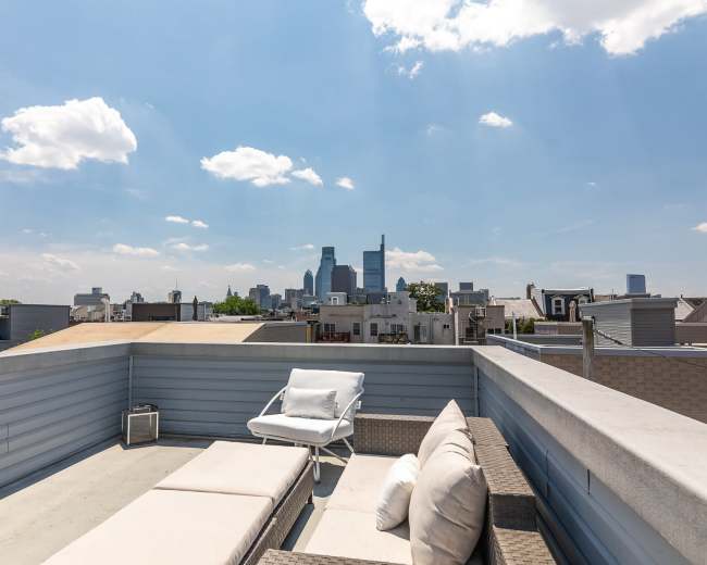 A rooftop terrace features lounge chairs overlooking a city skyline under a partly cloudy sky.