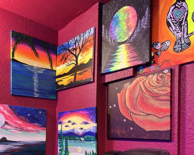 The image shows a restroom corner with a black counter, a soap dispenser, a paper towel dispenser, and several colorful paintings on the red walls.