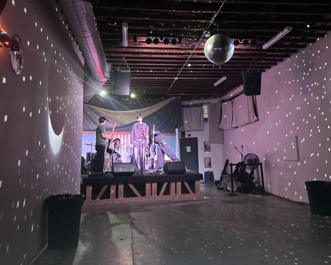 The image shows a performance space with a stage, drum set, and a person standing at the front, illuminated by colorful lights and a large disco ball reflecting patterns on the walls.
