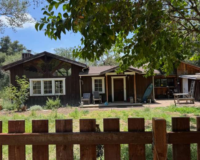 A wooden house with a porch, surrounded by greenery, is visible behind a wooden fence.