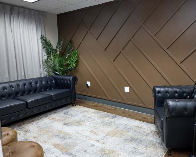 The image shows a room with two black leather tufted sofas facing each other, a light-colored patterned rug on wooden flooring, and a potted plant next to a brown paneled accent wall.