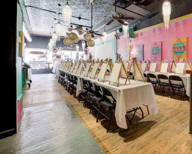 The image shows a spacious art studio set up for a painting event, featuring rows of empty easels and tables draped in white cloths.