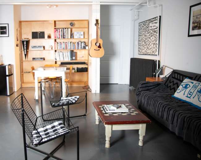 The image shows a modern living space featuring a black couch, a wooden table with chairs, bookshelves, and a guitar hanging on the wall.