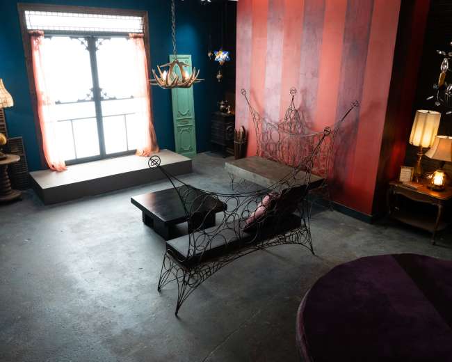 The image shows a stylish room featuring a metal wire sofa, a large round purple rug, and various lighting fixtures against a backdrop of colorful walls and a large window.