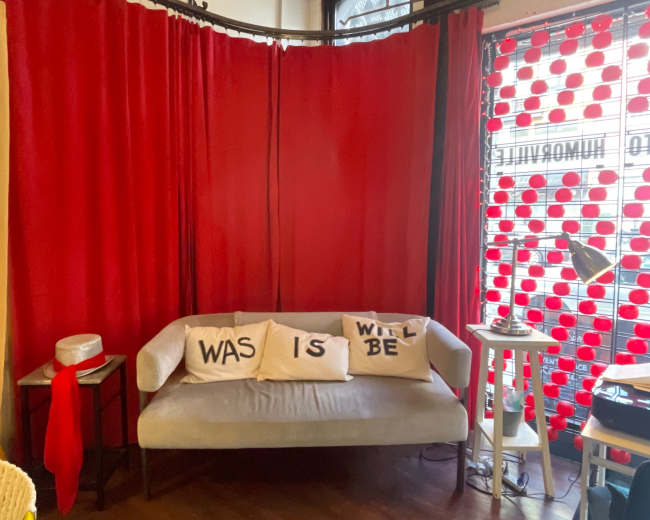 The image shows a cozy seating area featuring a light-colored sofa adorned with decorative pillows that read "WAS," "IS," and "WILL BE," against a backdrop of red curtains and a patterned window.