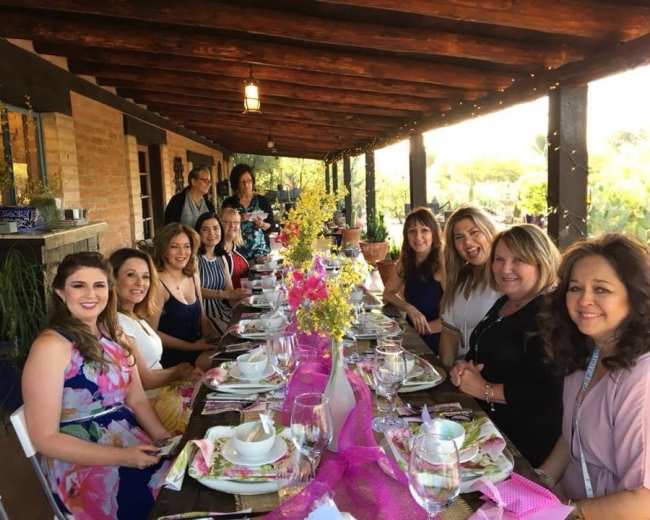 A group of women gathers around a long, decorated dining table on a porch, set for a meal in a rustic outdoor setting.