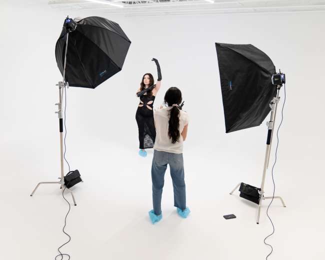 A model poses in a black outfit while a photographer captures the shot in a well-lit studio with two large softbox lights.