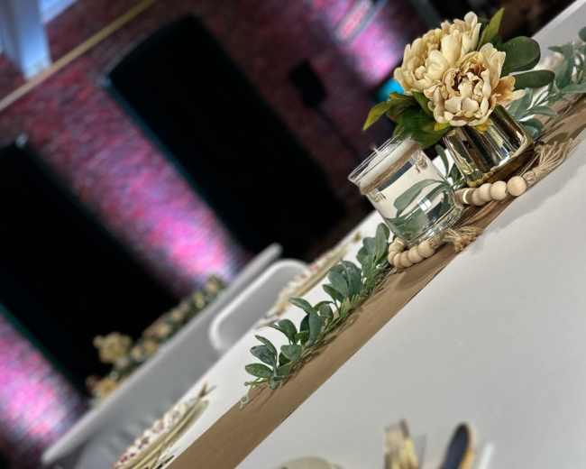 A dining setup featuring elegant tableware, a centerpiece with flowers, and decorative greenery on a table with a neutral table runner.