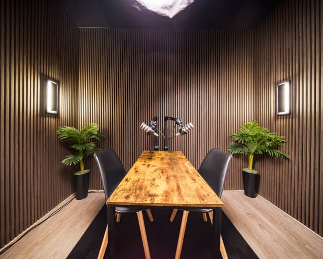 The image shows a modern podcasting studio with a wooden table, black chairs, and potted plants, surrounded by wooden paneling and soft lighting.