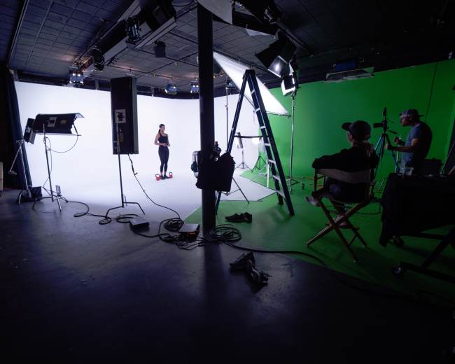 A photography studio is set up with two backgrounds, one white and one green, equipped with lights, a camera, a ladder, and two crew members observing a model on a stage.