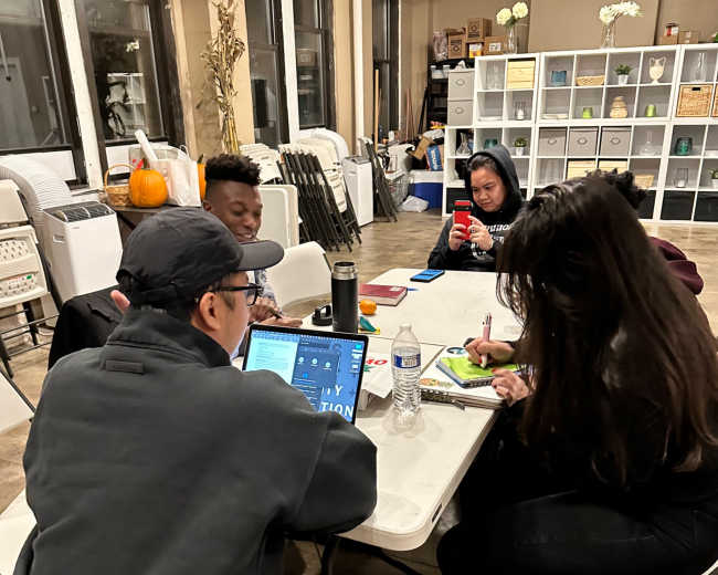 A group of four people collaborates around a table with laptops and notebooks in a well-lit indoor space.