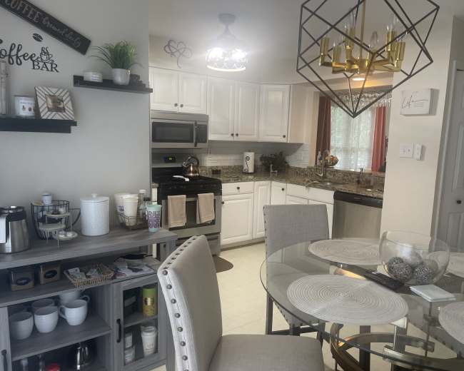 The image shows a modern kitchen with white cabinetry, granite countertops, and a dining area featuring a round table and elegant chairs.