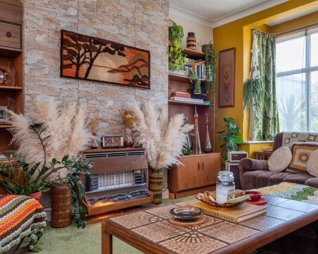 The image depicts a cozy living room featuring a stone accent wall, a brown sofa, decorative plants, and a coffee table adorned with books and a jar.
