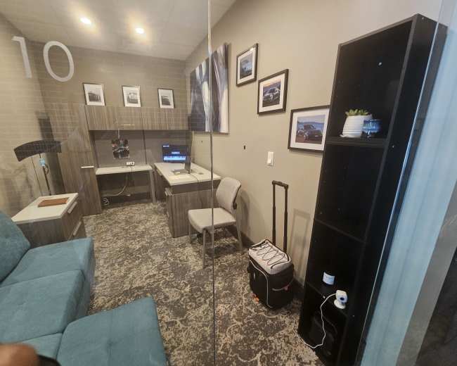 The image shows a small office room with a blue couch, a desk with a computer, a chair, a bookshelf, and a suitcase.