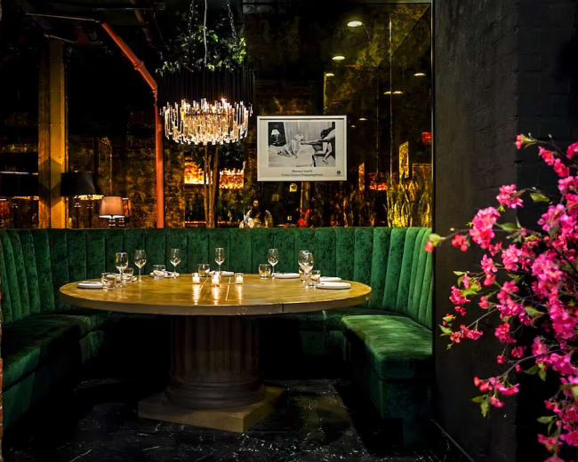 A round wooden table with place settings is centered in a green upholstered booth, surrounded by exposed brick walls and decorative elements like a chandelier and flowers.