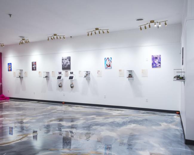 The image shows a modern art gallery with a polished floor, displaying various framed artworks on white walls and small shelves.