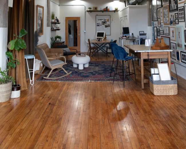 The image depicts an open, modern workspace featuring wooden floors, a ceiling fan, and various seating and work areas arranged around framed photographs on the walls.