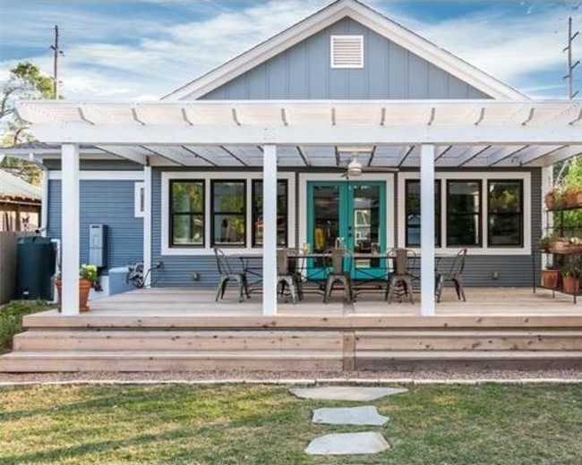 The image shows a blue house with a large wooden deck featuring a covered patio and a set of chairs and tables.