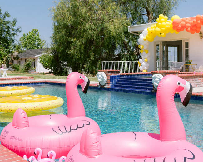 Two pink flamingo-shaped pool floats are positioned in a swimming pool beside a brick edge, with colorful balloons decorating an area in the background.