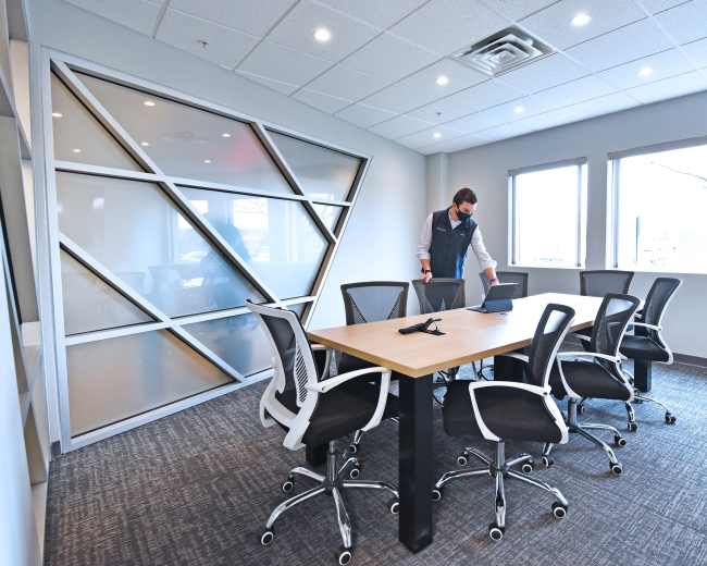 A person is preparing a conference room with a large table and several ergonomic chairs, while standing near a laptop.