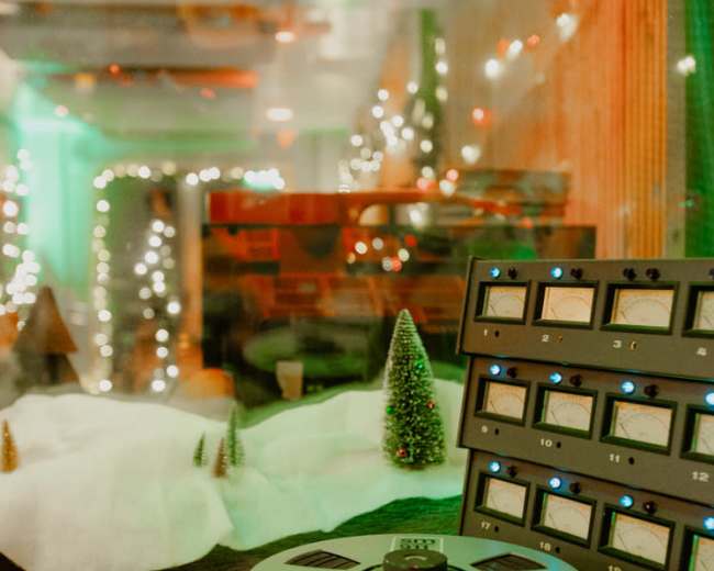 The image shows a recording studio setup with a vintage reel-to-reel tape machine and a control panel displaying various gauges, surrounded by festive decorations and miniature trees.