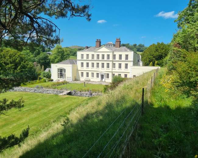 A large, yellow-painted house stands on a hillside, surrounded by a green lawn and trees under a blue sky.