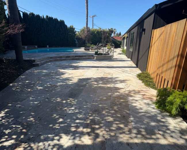 The image shows a stone-paved outdoor area next to a swimming pool, with palm trees and a modern building in the background.