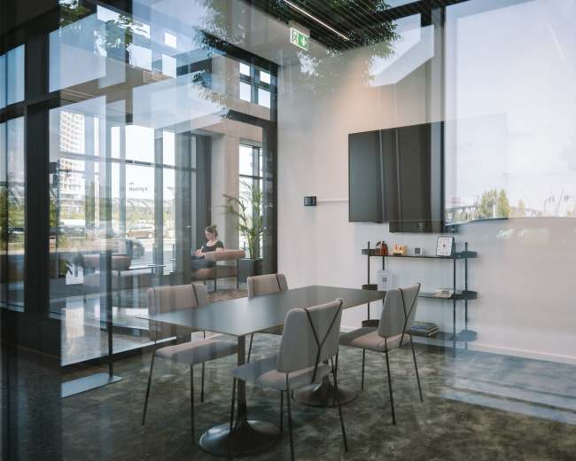 A modern conference room features a large table surrounded by chairs, with a television mounted on the wall and large windows providing a view of the exterior.