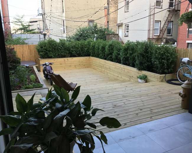 The image shows a wooden deck surrounded by greenery, with a vintage scooter and a bicycle parked nearby.