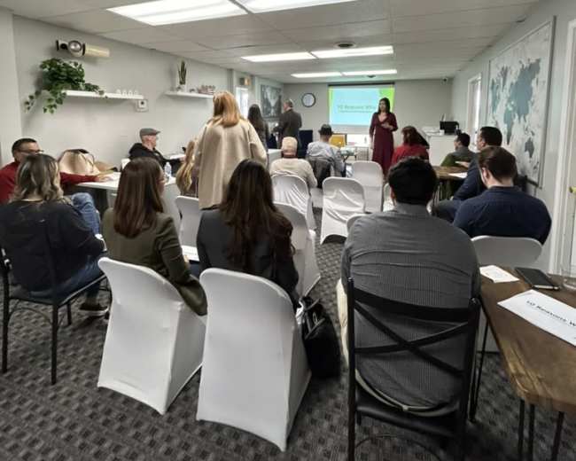 A group of people is seated in a conference room, watching a presentation displayed on a screen at the front.