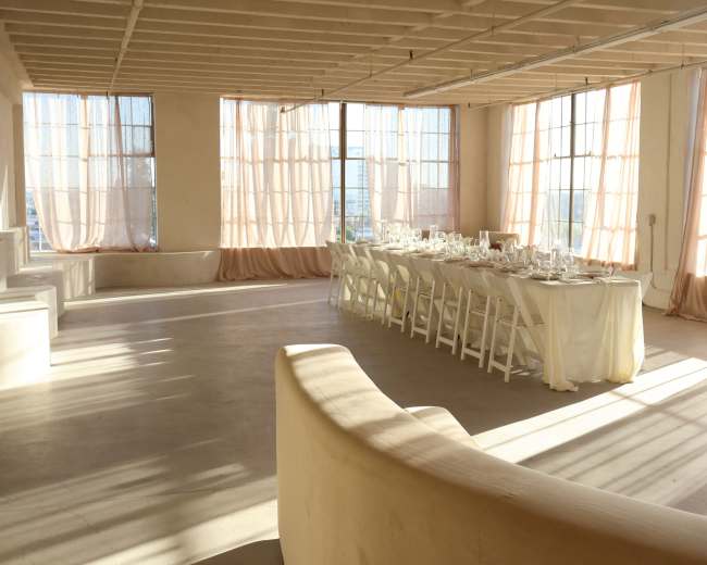 A long dining table is set with glassware and white table linens in a spacious, well-lit room with large windows and soft drapes.