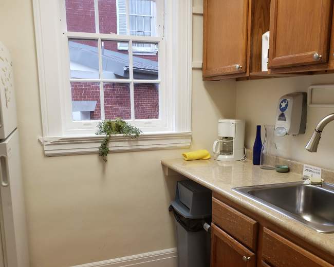 The image shows a small kitchen area with wooden cabinets, a sink, a coffee maker, and a window partially revealing a brick building outside.