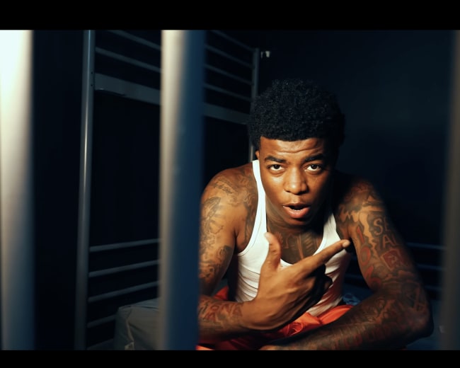 A man with tattoos sits on a bed in a cell, gesturing toward the camera while looking directly at it.