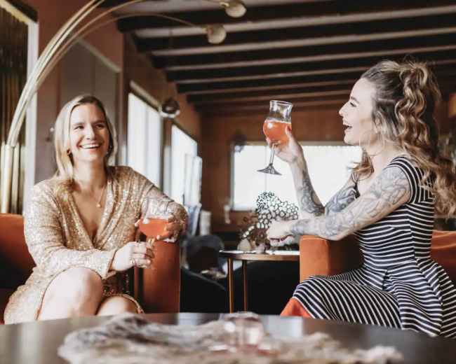 Two women sit on orange couches, holding drinks and smiling at each other in a well-decorated indoor space.