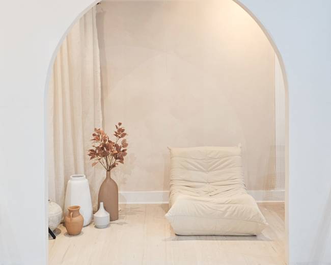 A minimalist interior features a padded chair beside vases and dried foliage, framed by a curved archway.