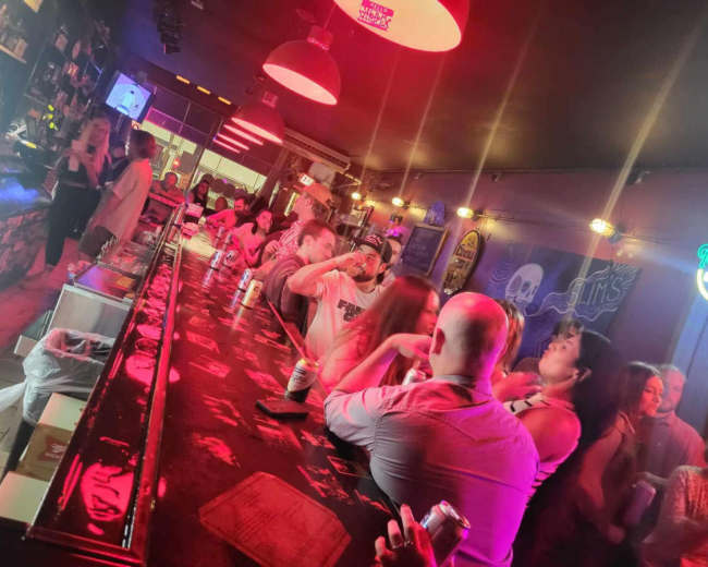 A crowded bar scene features a long counter with patrons socializing under dim red lighting.