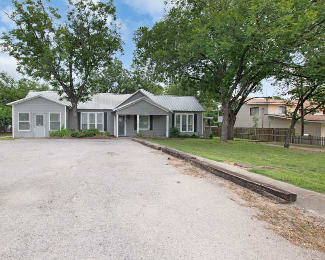The image shows a single-story house with a metal roof surrounded by trees and a gravel driveway.