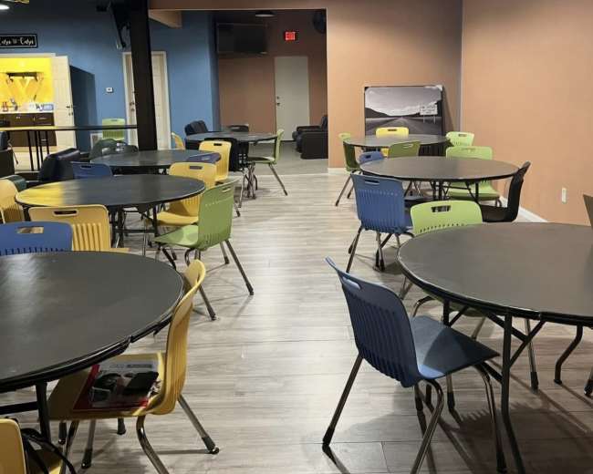 The image shows a brightly lit room with multiple round tables and chairs in various colors arranged throughout the space.