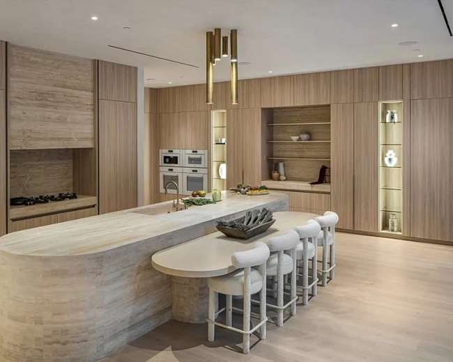 The image depicts a modern kitchen featuring a curved stone island with seating and wooden cabinets, complemented by built-in appliances and ambient lighting.