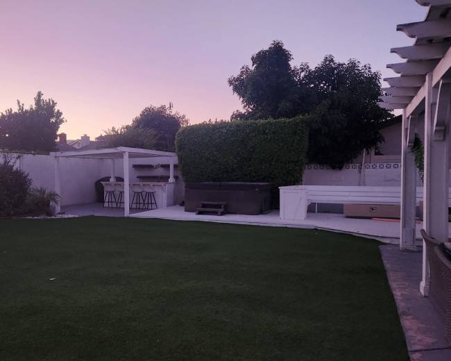 The image shows a backyard with artificial grass, a covered outdoor kitchen area, and trees against a pastel-colored sky at dusk.