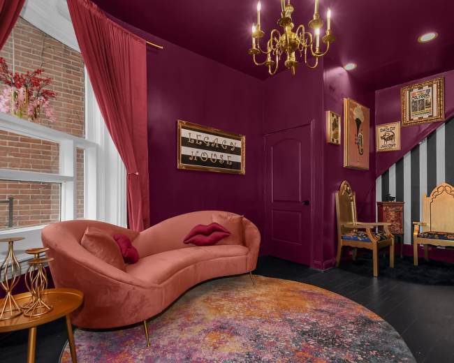A richly colored room with a curved pink couch, a round rug, a chandelier, and decorative wall frames, featuring a staircase in the background.