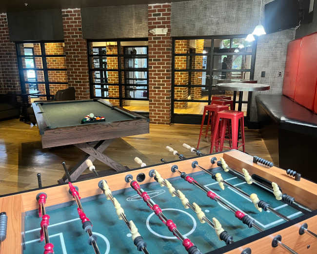 The image shows a recreational area featuring a foosball table in the foreground, a pool table in the background, and brick walls with large glass doors.