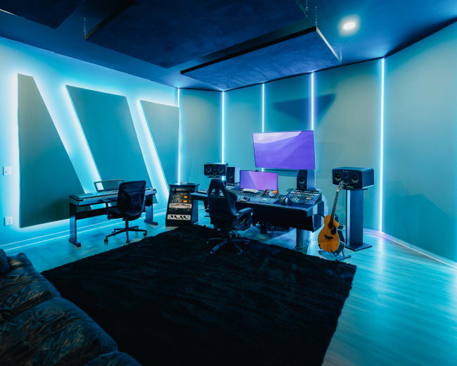 The image shows a modern music studio equipped with a keyboard, audio monitors, a mixing desk, and ambient lighting along the walls.